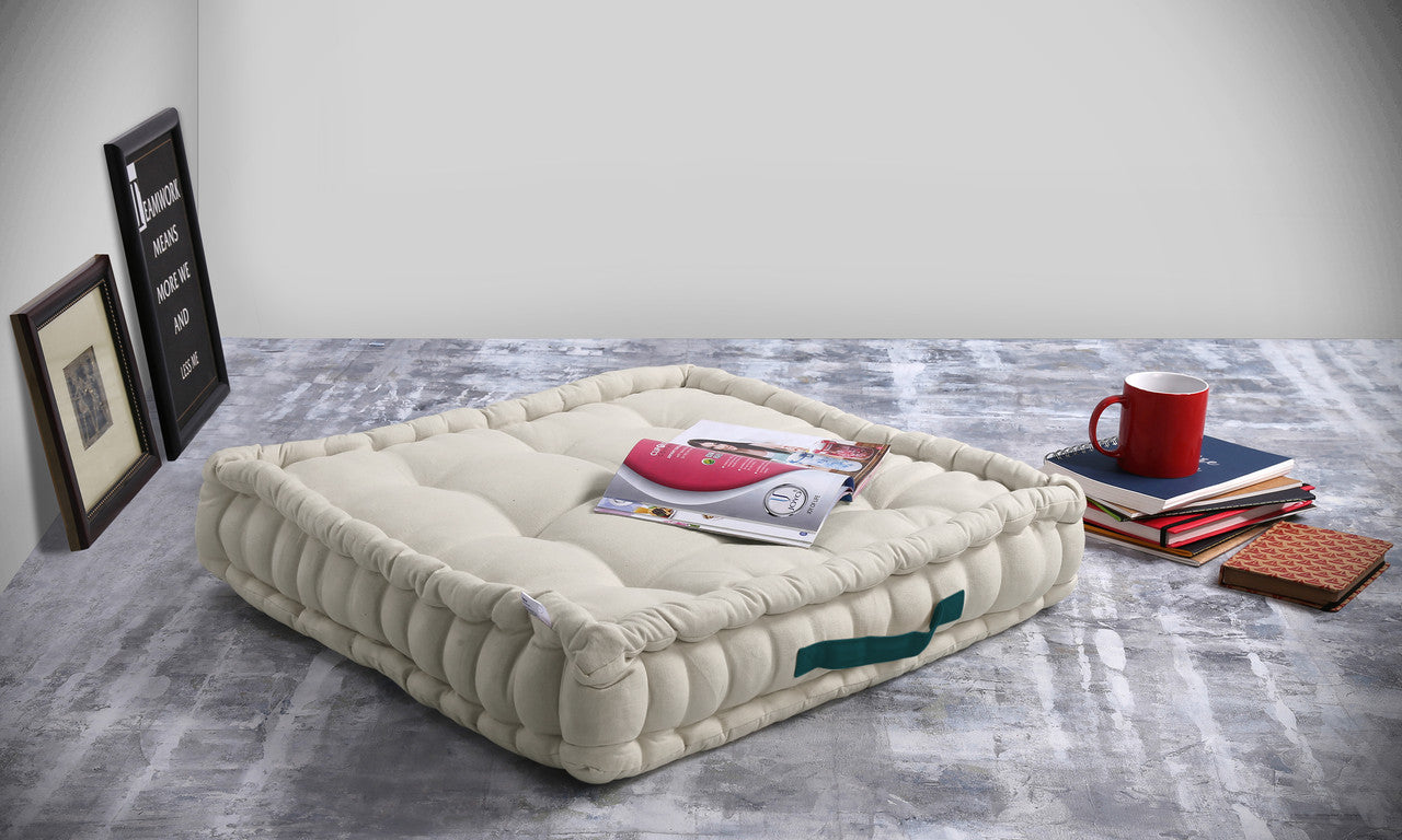 Off-white square cotton floor cushion with a teal handle on a grey floor, open magazine on it, red mug on books