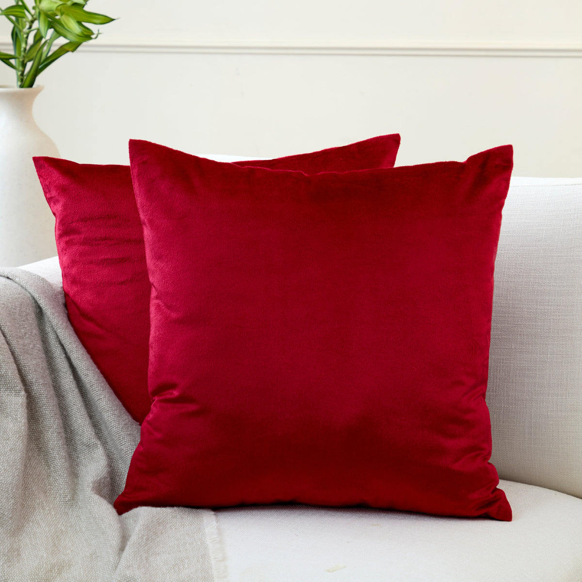 Two red velvet cushion covers stacked on an off-white sofa beside a plain grey throw