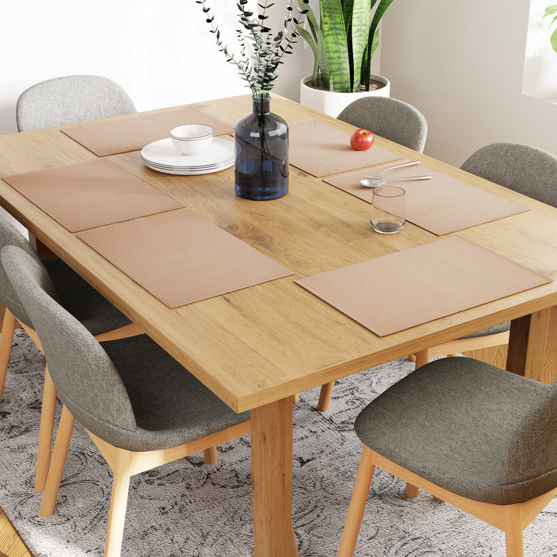 Six ribbed beige placemats on a bare oak dining table with stacked white plates, a blue glass vase and one red apple